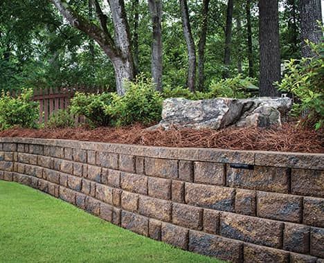 Retaining wall made of brown blocks with a green lawn in the foreground and trees in the background.