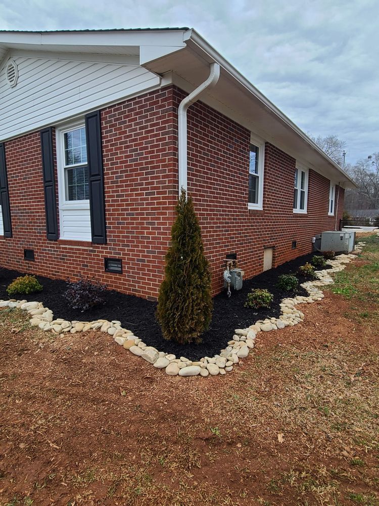 Red brick house corner with new landscaping, black mulch, small shrubs, and river rock edging.