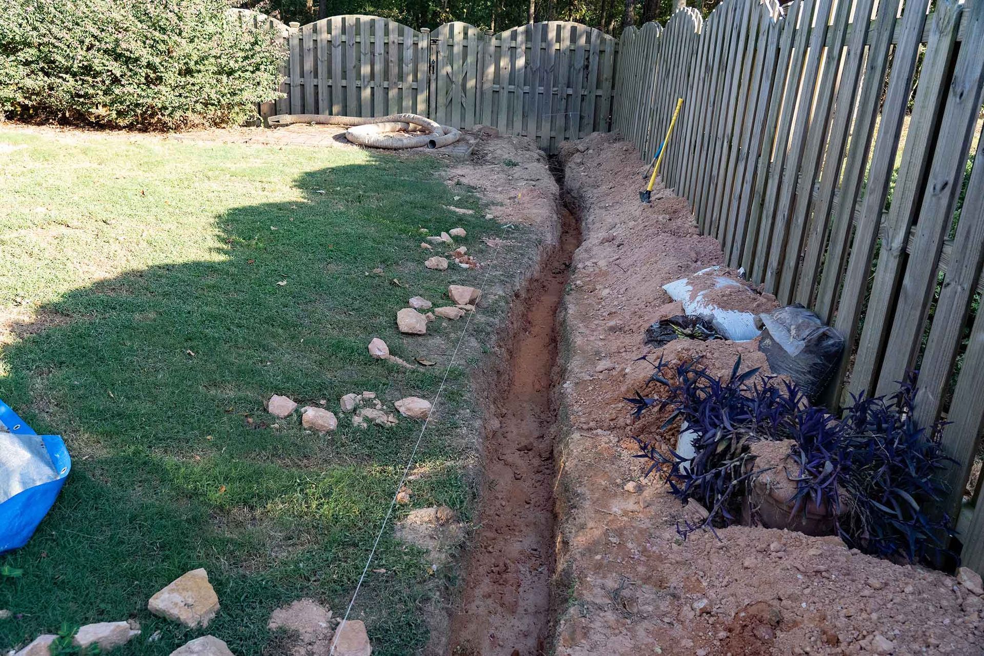 A narrow trench dug in a backyard, beside a wooden fence, running through grass.