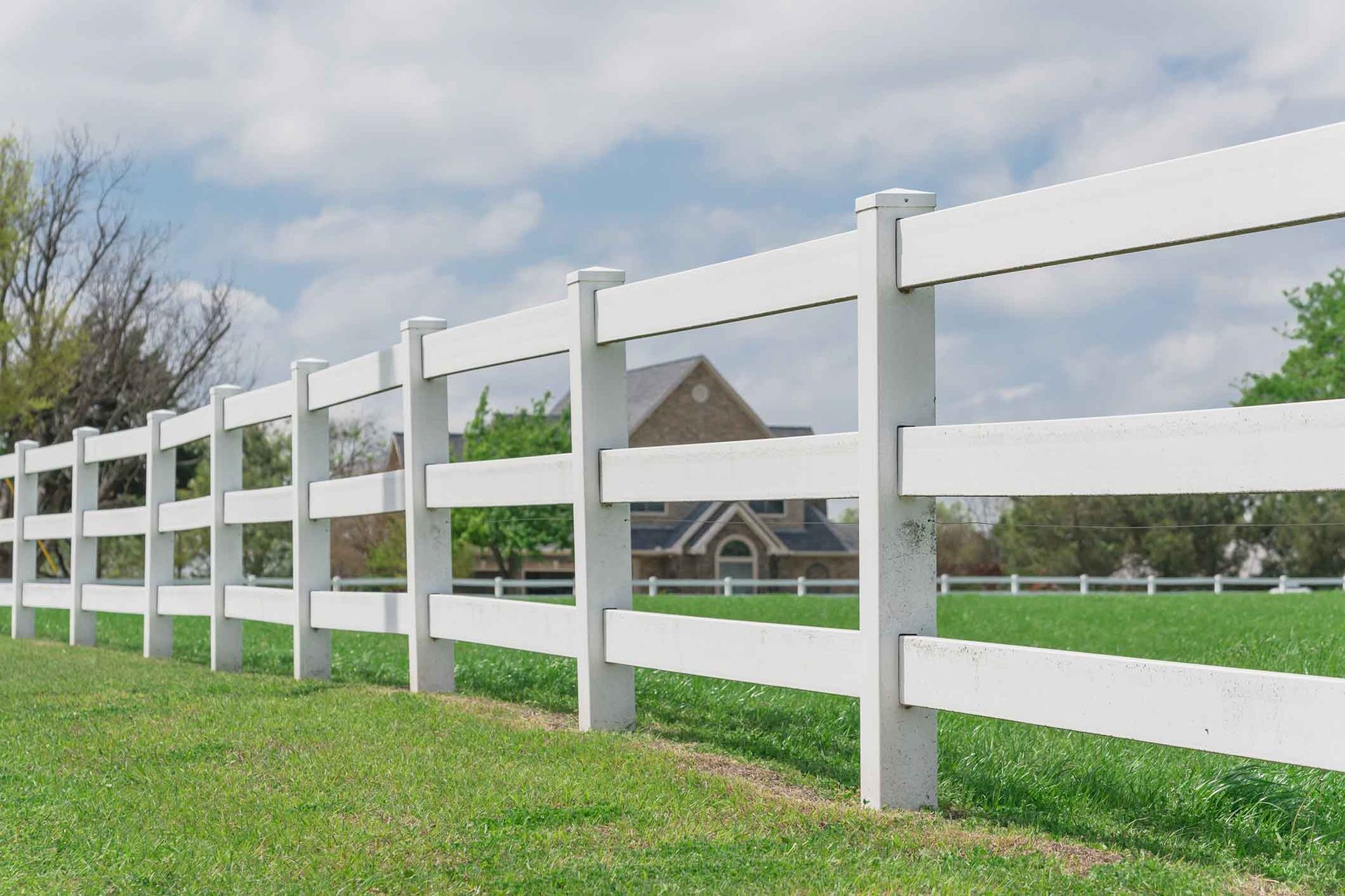 White vinyl fence in a green field with a house in the background and a partly cloudy sky.