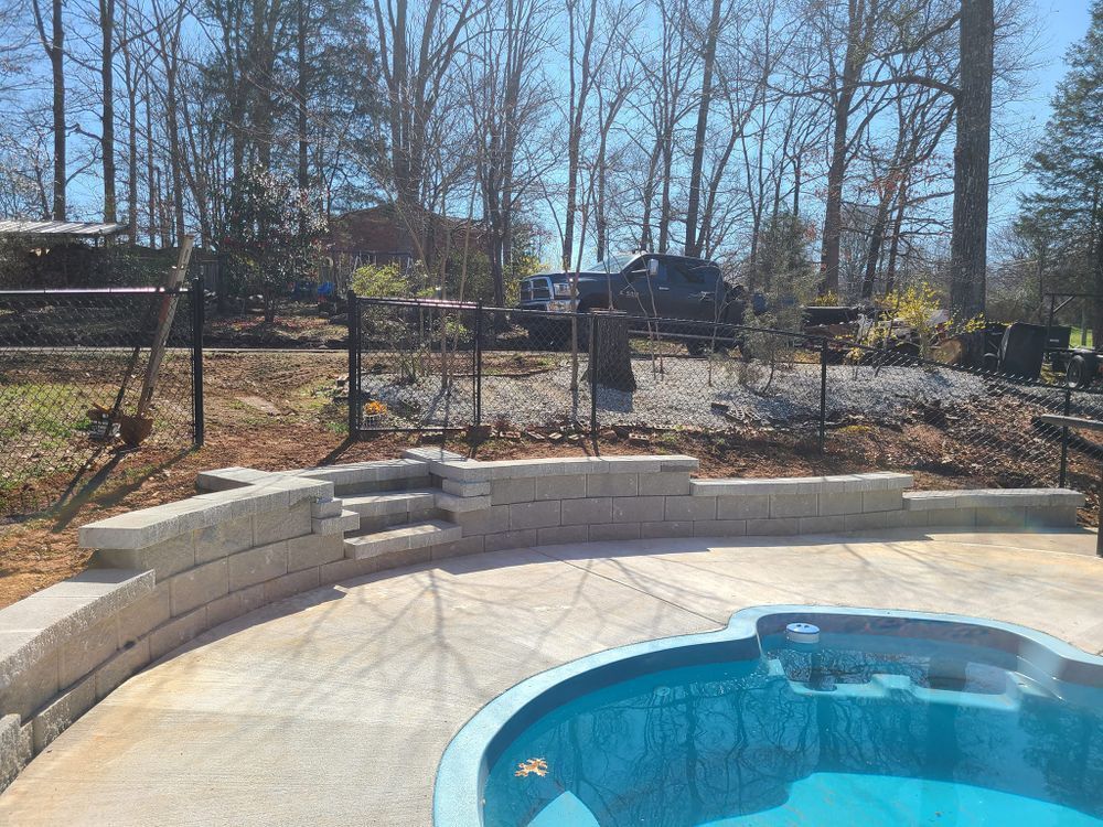 A swimming pool and patio area with retaining walls and a black fence in a wooded setting.