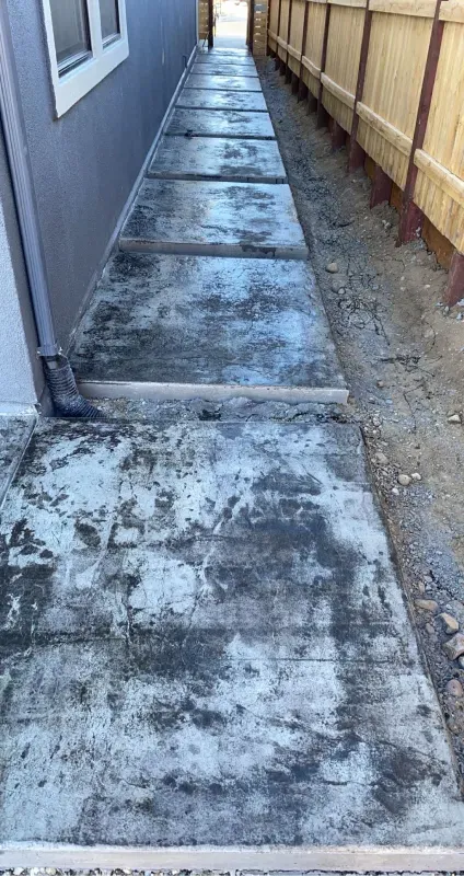 A concrete walkway leading to a house next to a wooden fence.