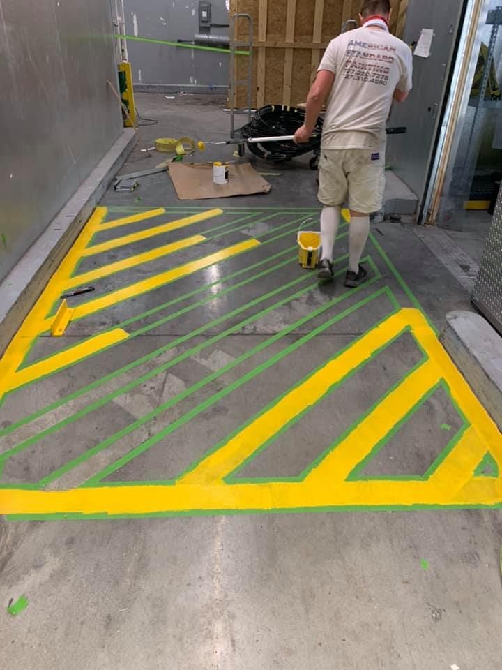 Person painting yellow hazard lines on a concrete floor, using green tape as a guide, indoors.