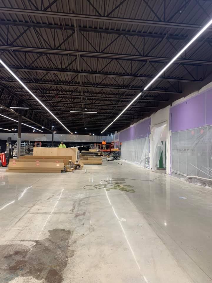 Construction in a large retail space with fluorescent lighting. Exposed ceiling, concrete floor, purple walls, and building materials.