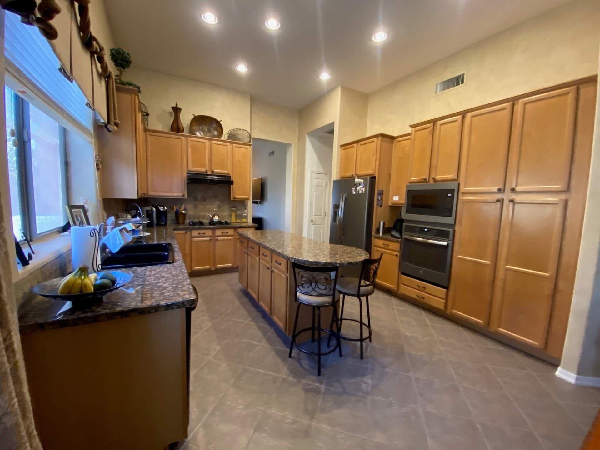 Spacious kitchen with wooden cabinets, island with stools, granite countertops, stainless steel appliances, and tiled floors.
