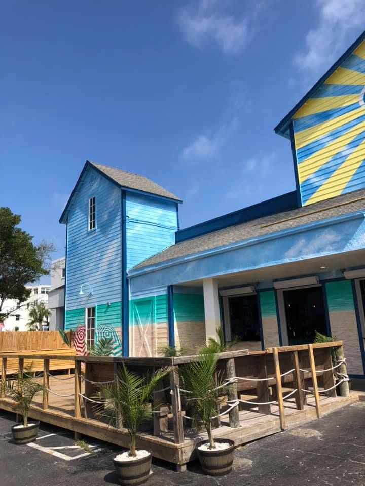 Brightly painted beachside building with a deck, featuring blue, yellow, and sand-colored walls against a clear sky.
