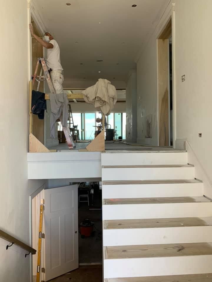 Person painting a doorway, standing on a ladder, with stairs in the foreground and a hallway in the background.