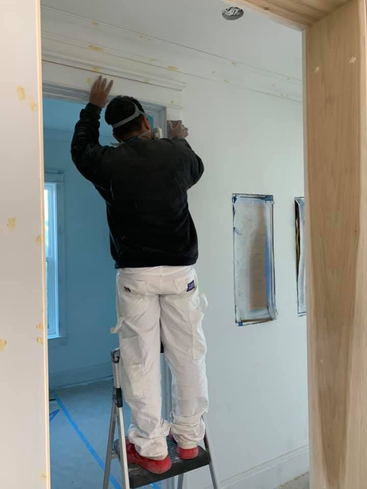 A person, wearing a mask, paints crown molding while standing on a ladder in a room.