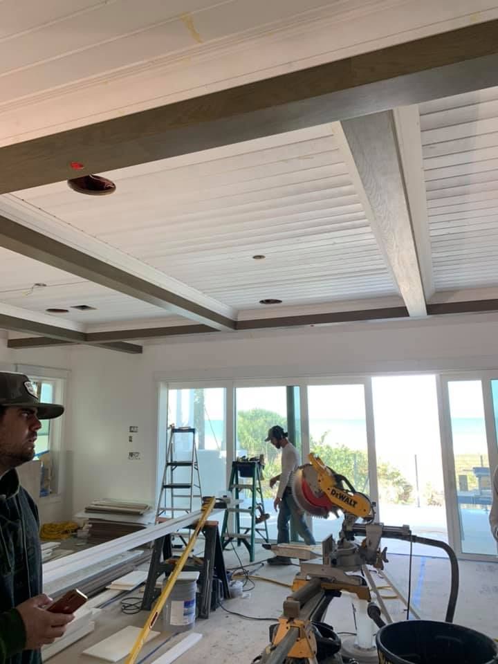 Construction in progress: men installing ceiling beams and trim in a sunlit room with ocean view.