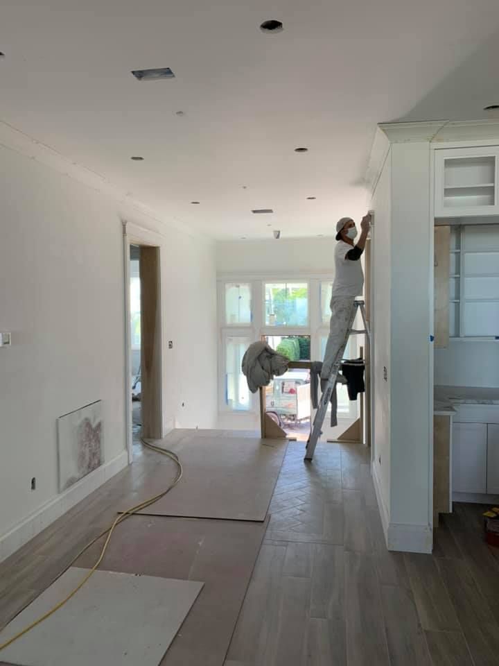 Person on a ladder painting a white wall in a room with wooden floors.