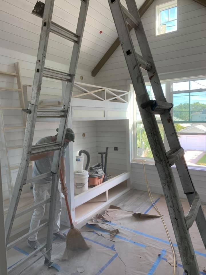 Person sweeping dust in a room with ladders and white walls; construction in progress.