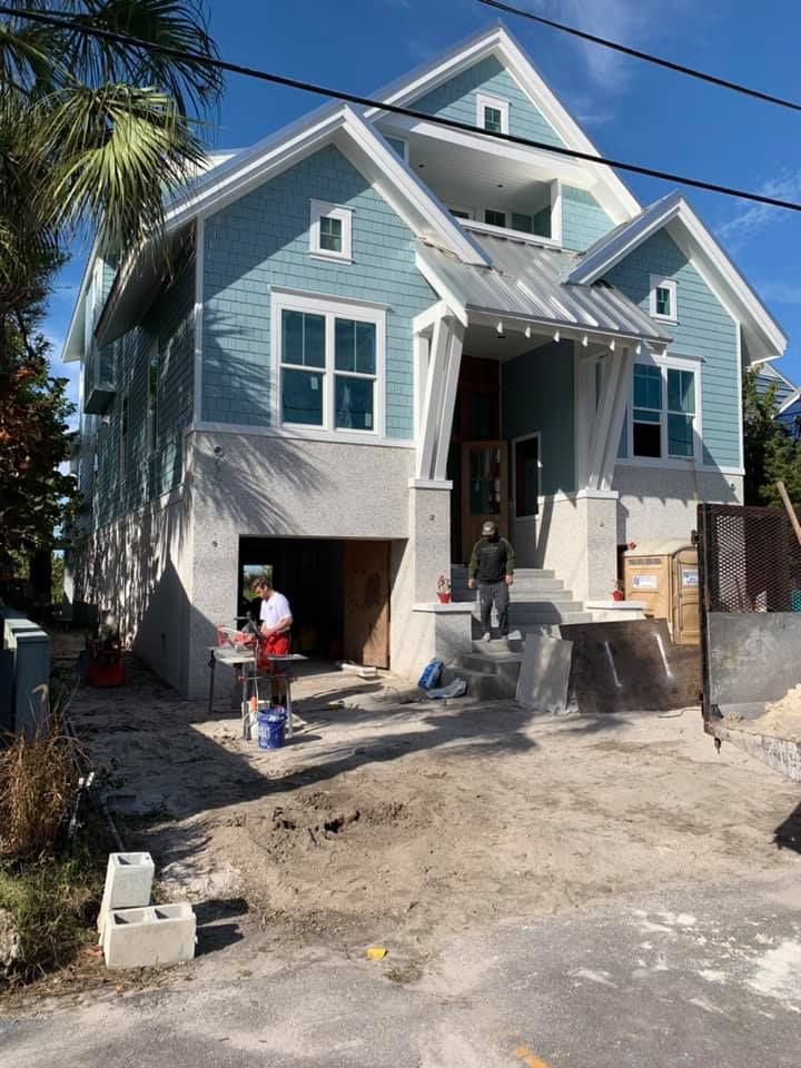 Three-story house under construction with blue siding, two people present, and a sandy driveway.