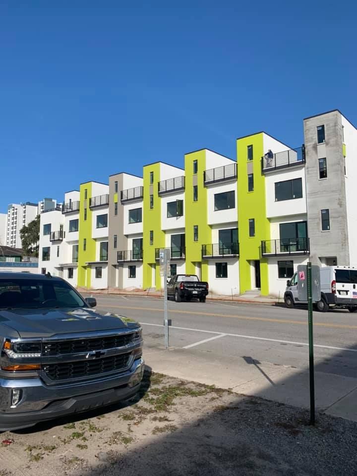 Modern row houses with green and white facades on a sunny street, with cars parked in front.
