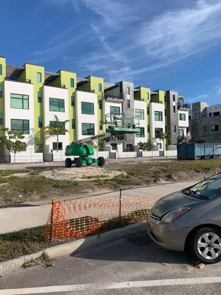 Green and white townhouses with a lift, car and construction fencing on a sunny day.