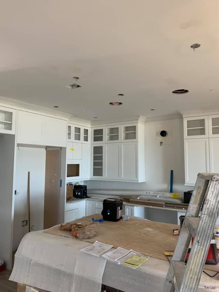 White kitchen cabinets installed, construction in progress. Island with items, ladder, ceiling fixtures.
