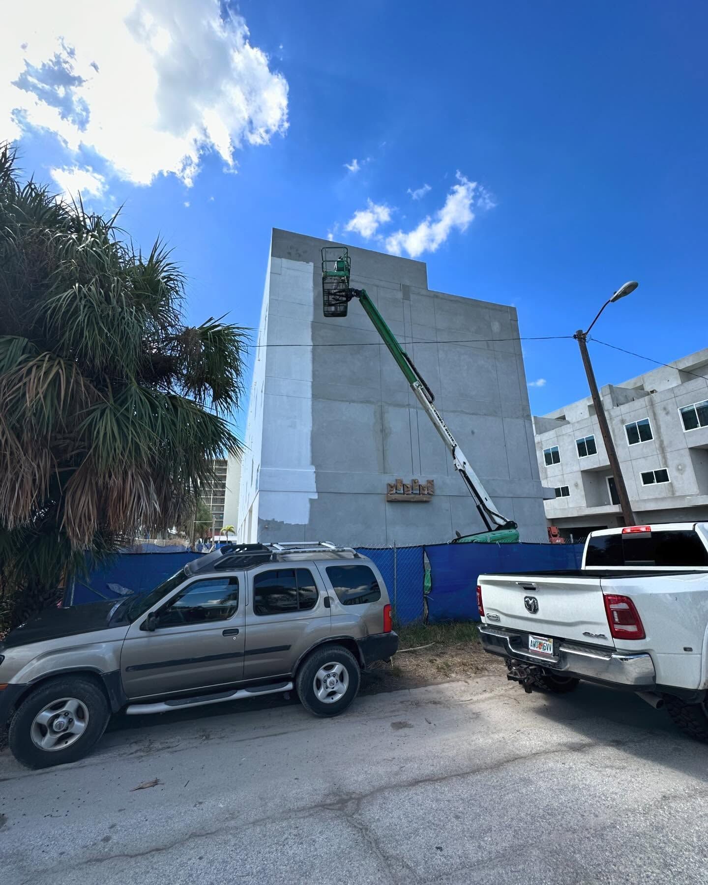 Construction site with a lift painting a gray building, blue fence, cars parked nearby, blue sky.
