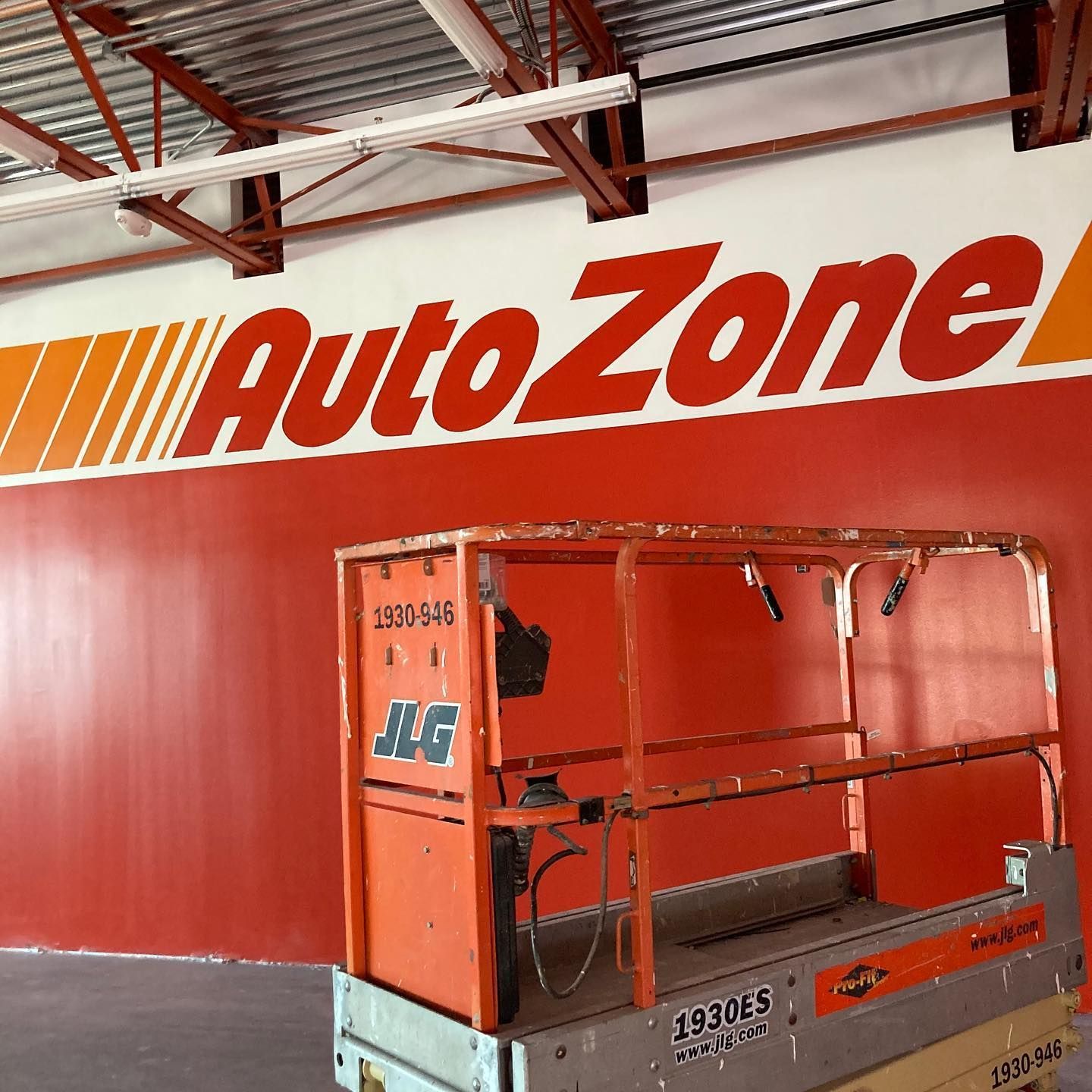 An orange JLG lift in an AutoZone store under the company's logo on a red wall.