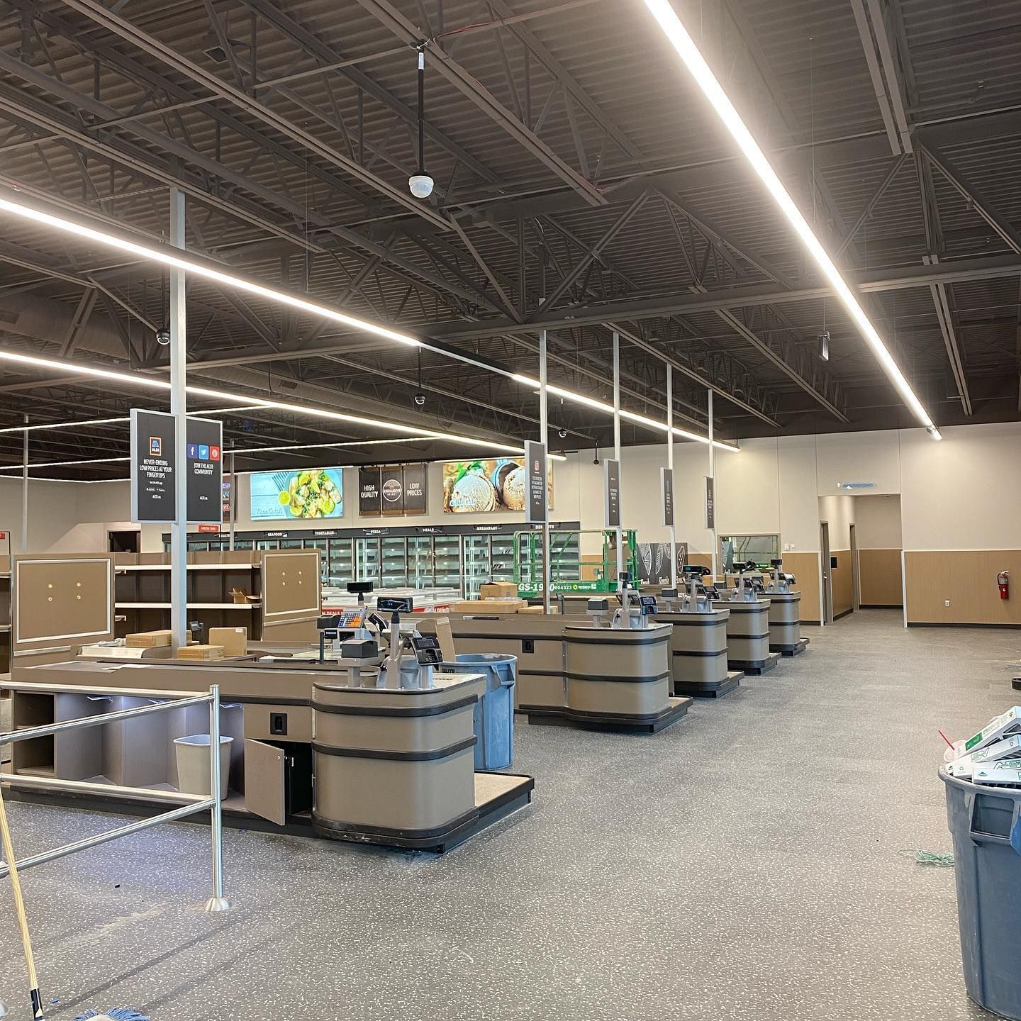 Empty grocery store checkout lanes, partially constructed. Gray floors, brown counters, white overhead lights.