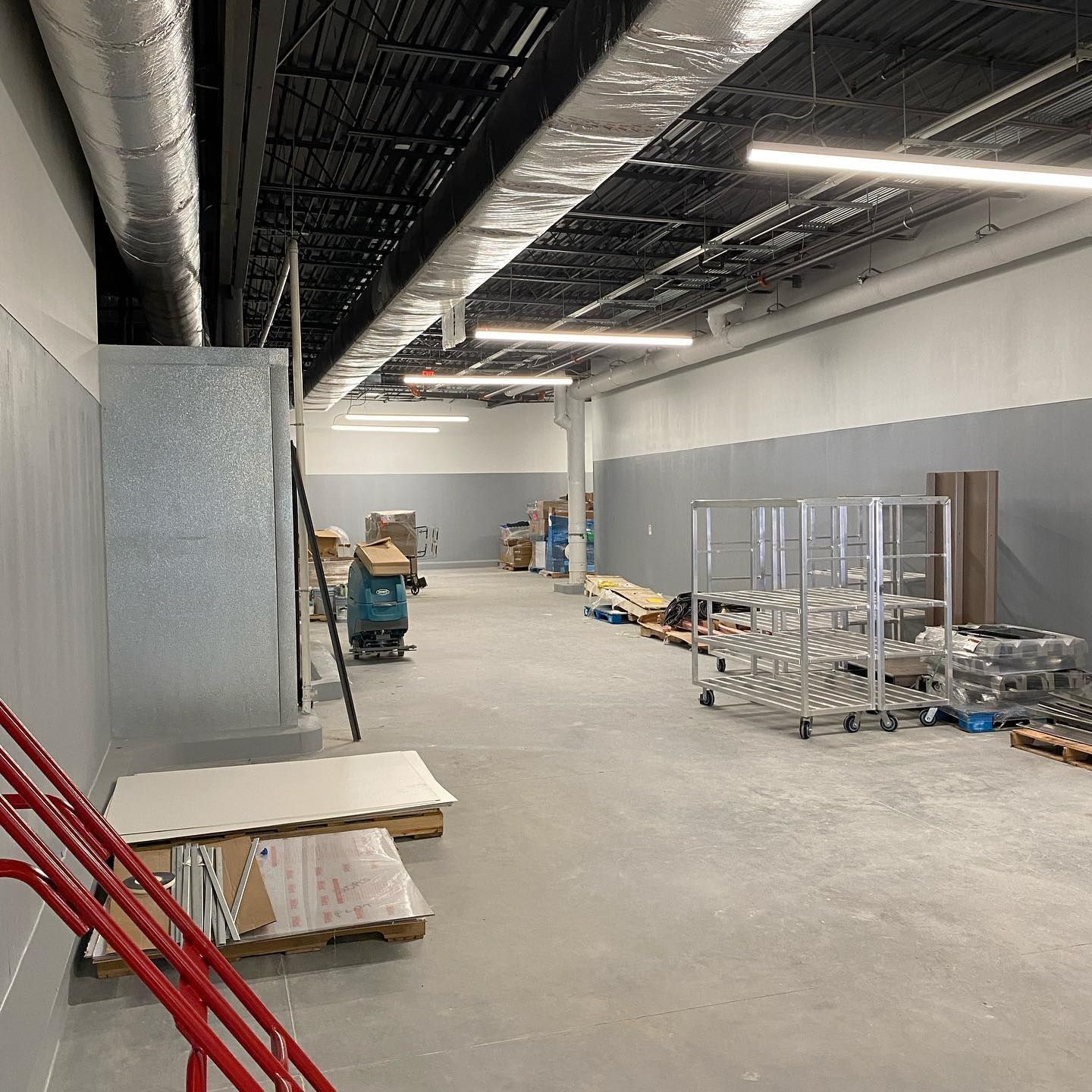 Interior of a large warehouse with exposed ductwork and long fluorescent lights, construction materials on the floor.