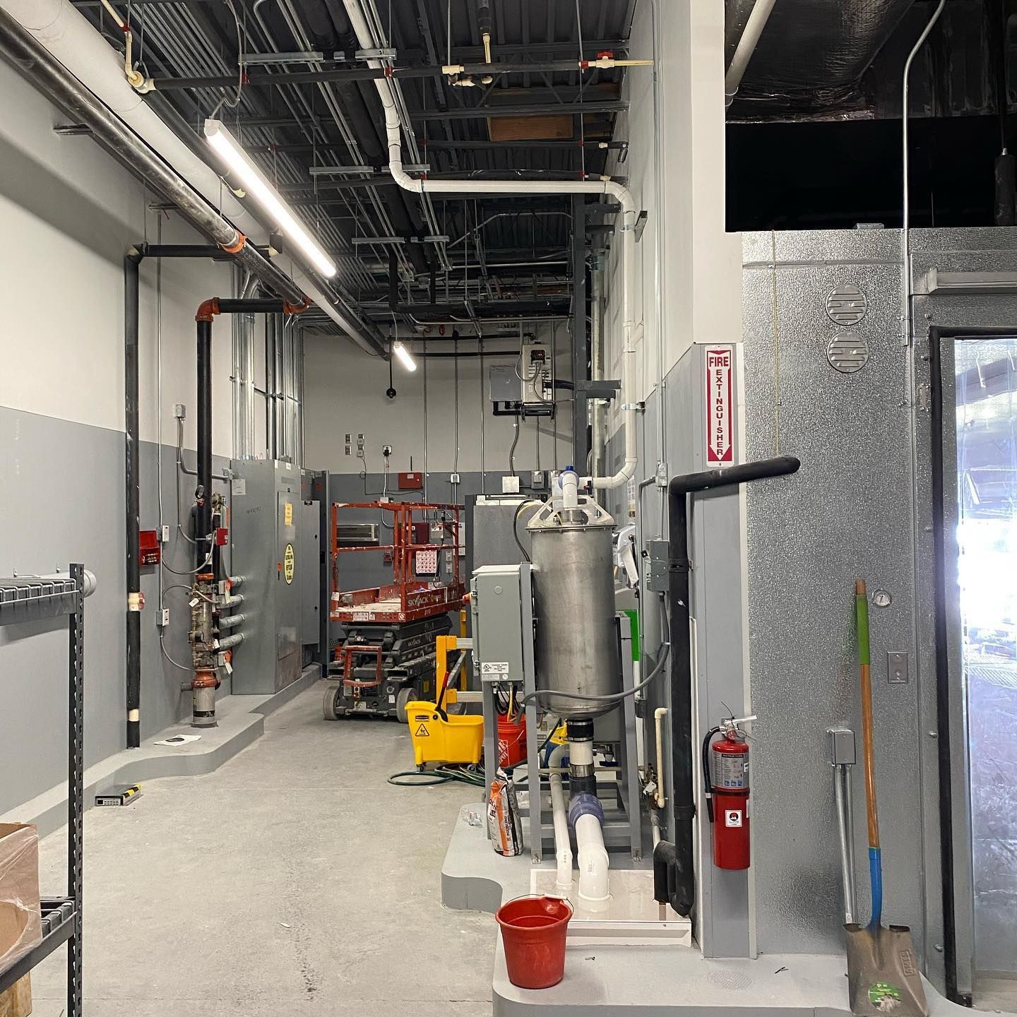 Industrial room with pipes, tanks, and equipment; gray and white walls, fire extinguisher.