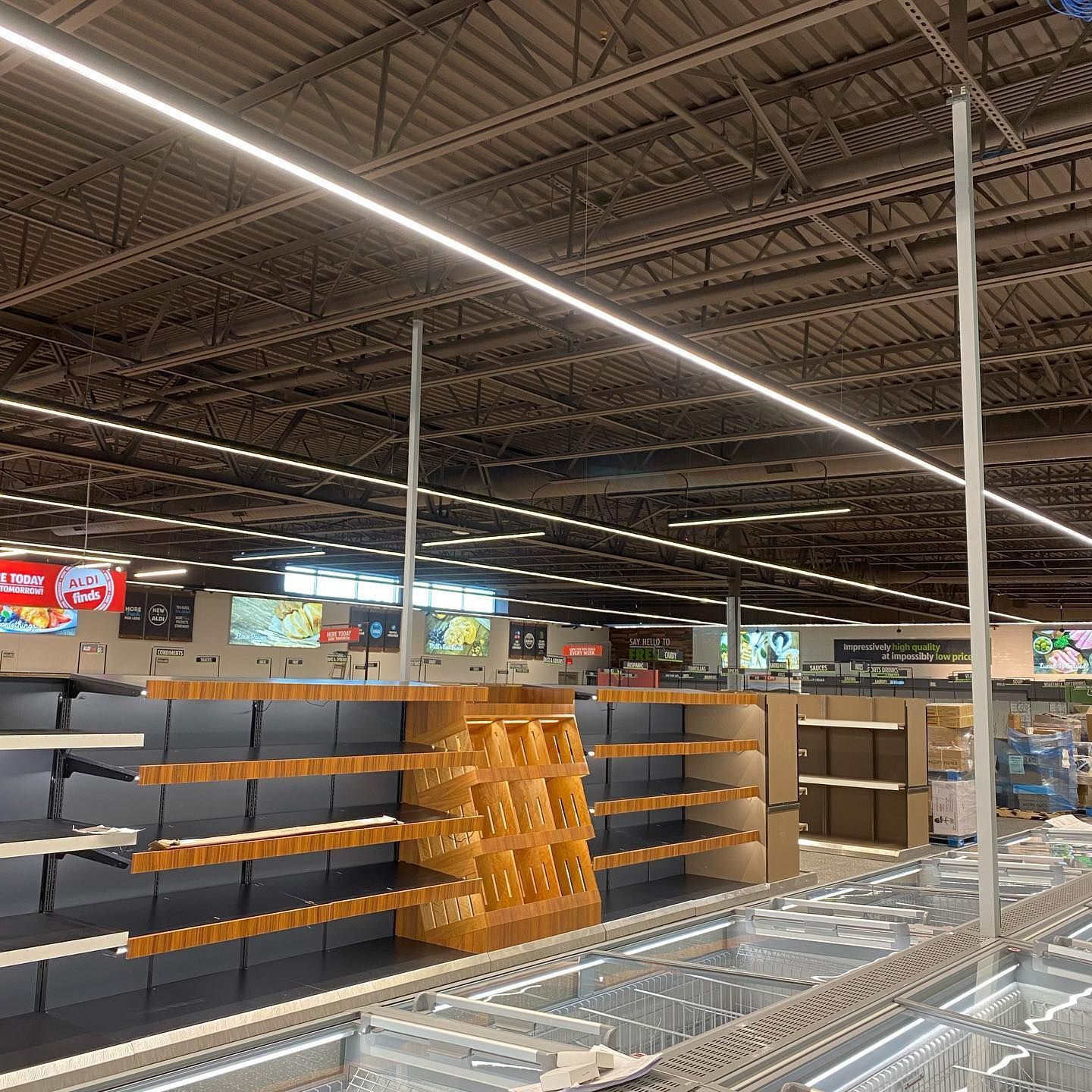 Empty grocery store shelves, overhead lighting, partially stocked middle display, dark ceiling.