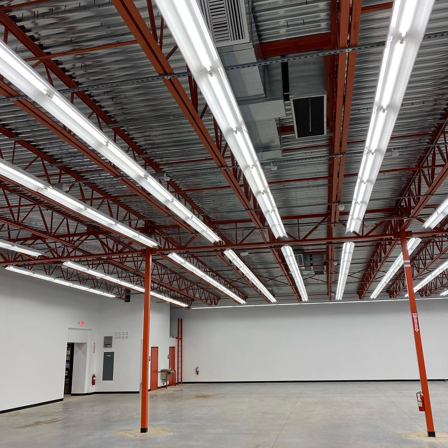 Empty industrial space with red beams, concrete floor, and fluorescent lights. White walls.