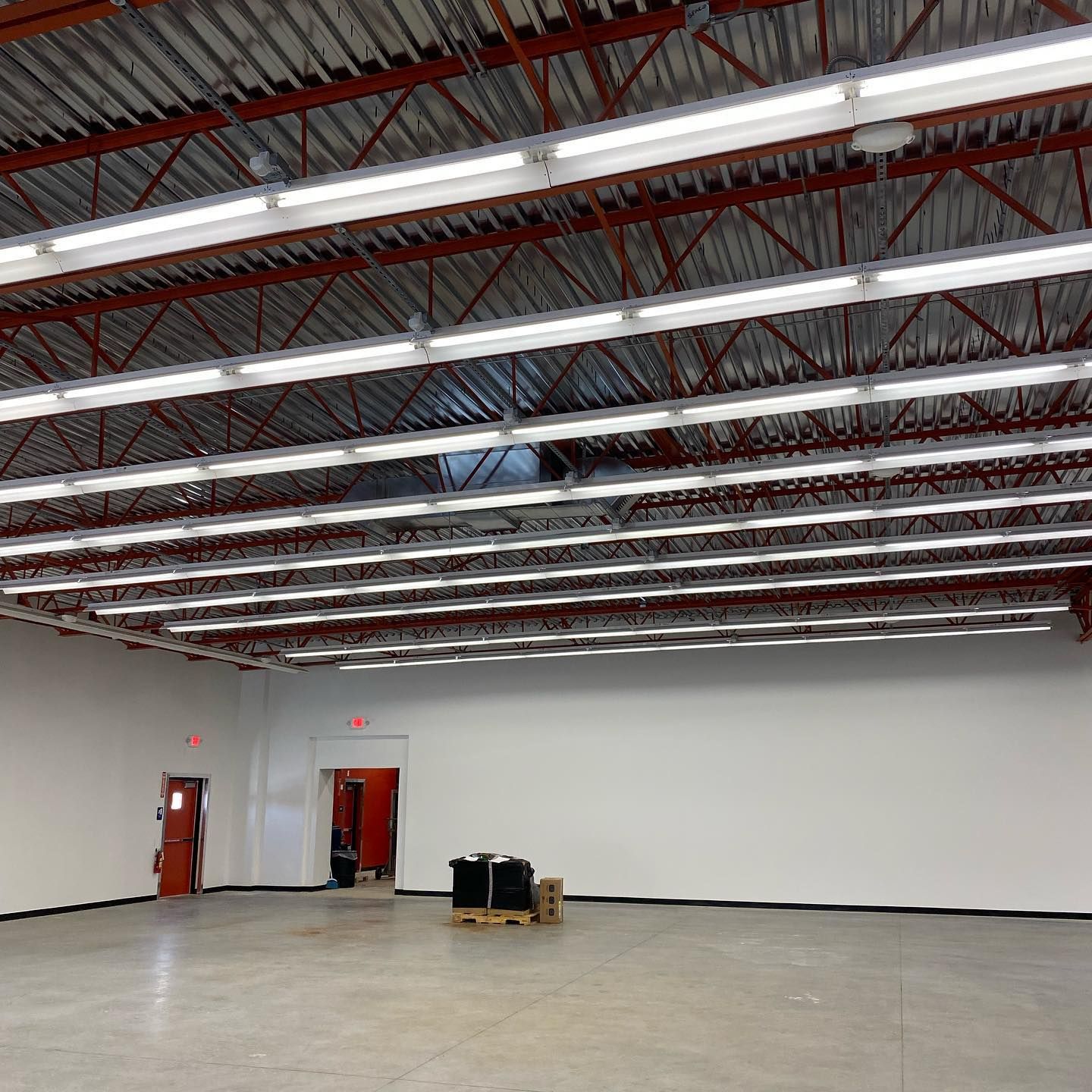 Empty warehouse interior with white walls, red metal trusses, and overhead fluorescent lights.
