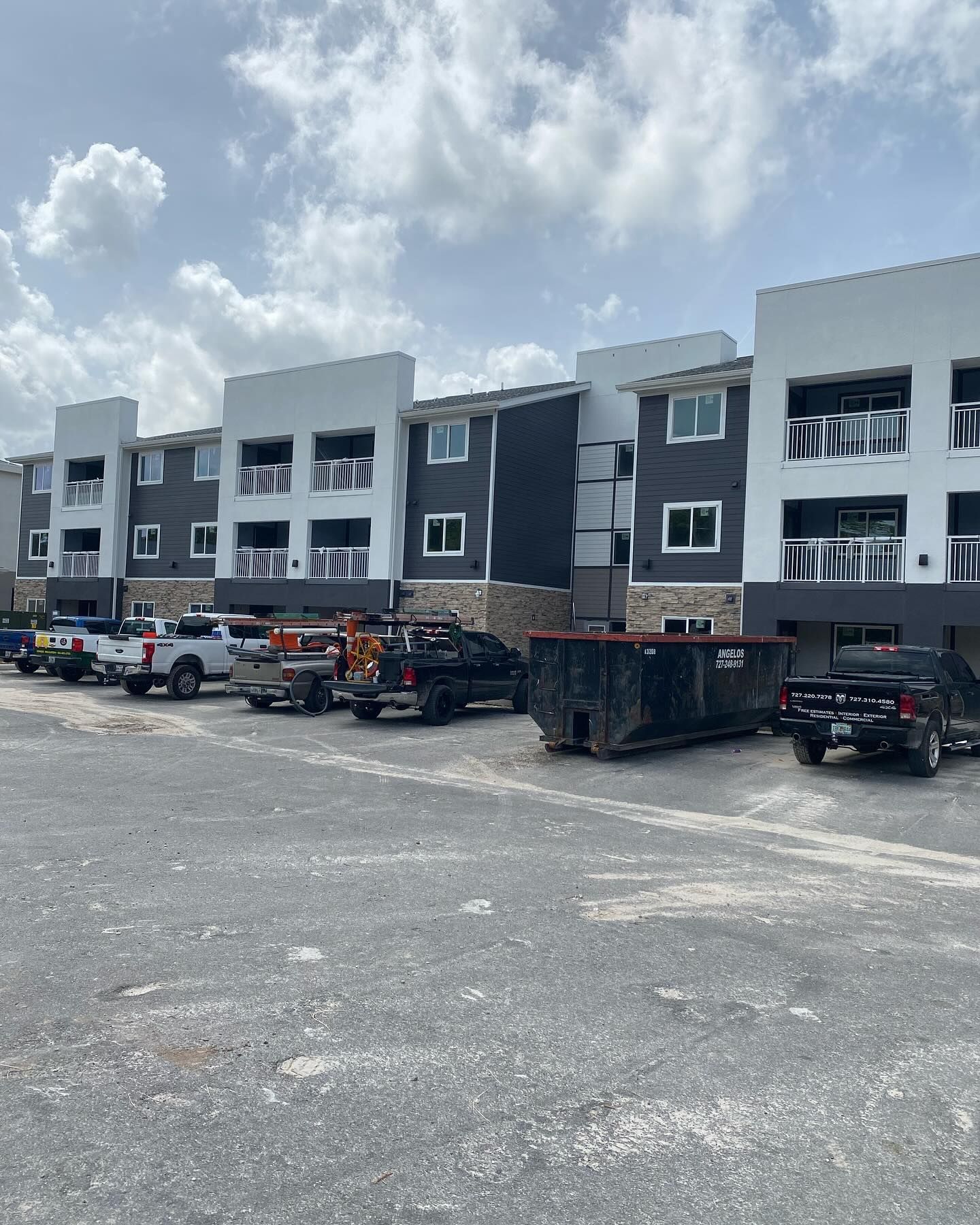 Construction site with multi-unit apartments, gray and white facade, vehicles and dumpster on a gravel lot.