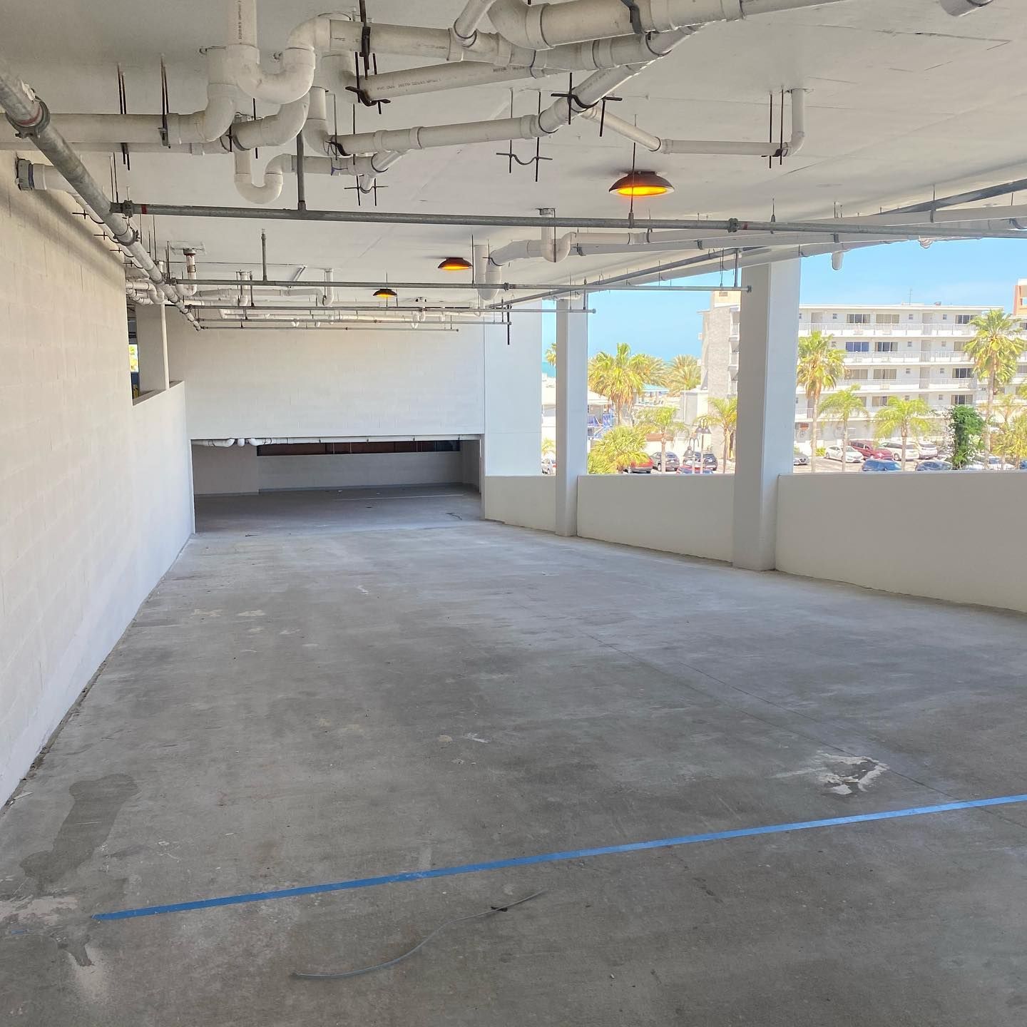 Empty parking garage with white walls, pipes, and a view of palm trees and buildings.