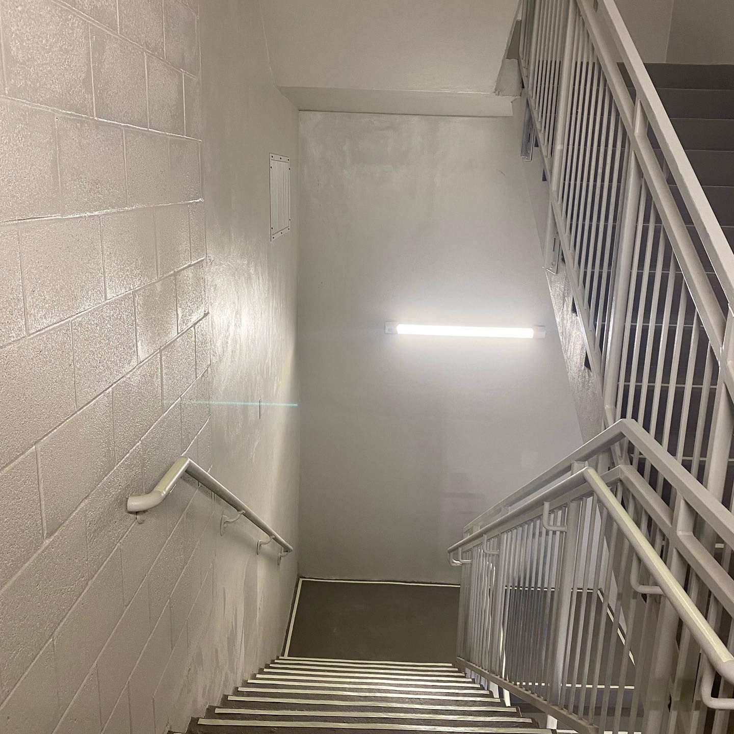 Stairwell with gray stairs and white walls, handrails. Fluorescent light fixture.