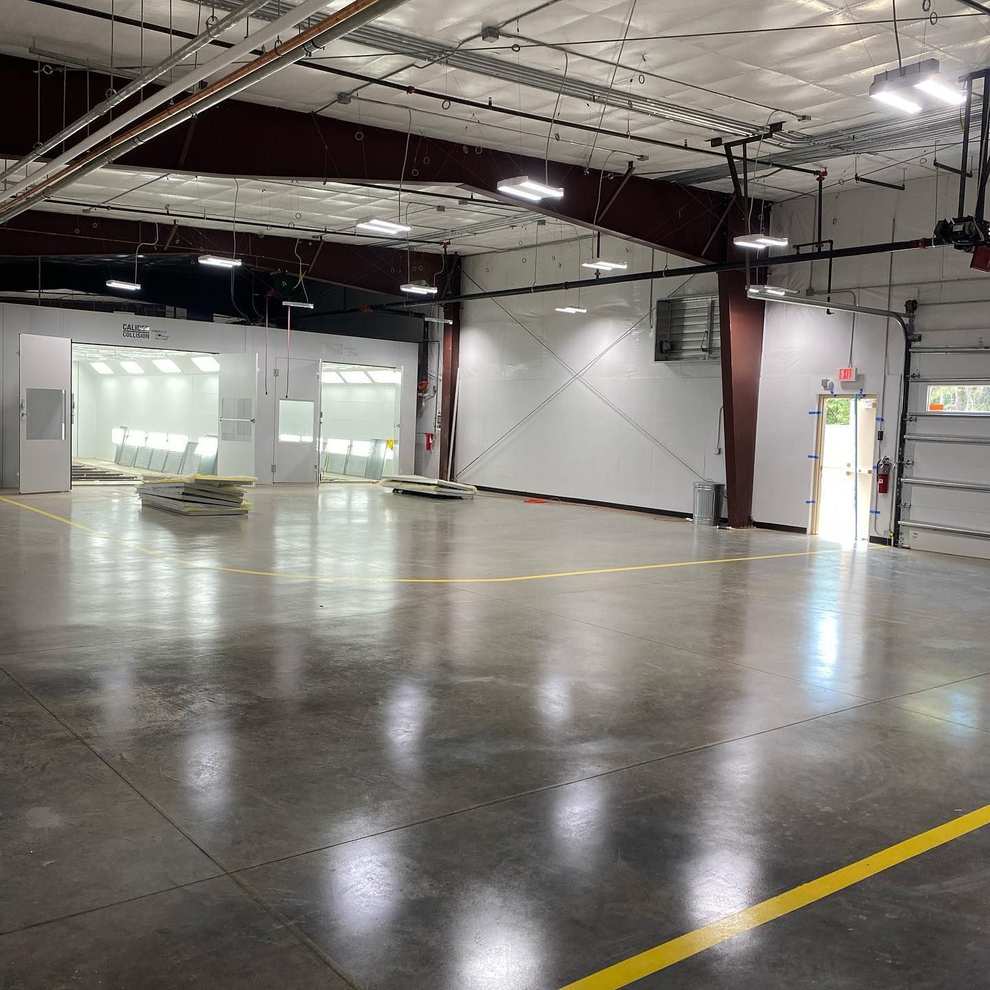 Large, empty industrial space with polished concrete floor, spray paint booth, and roll-up door.