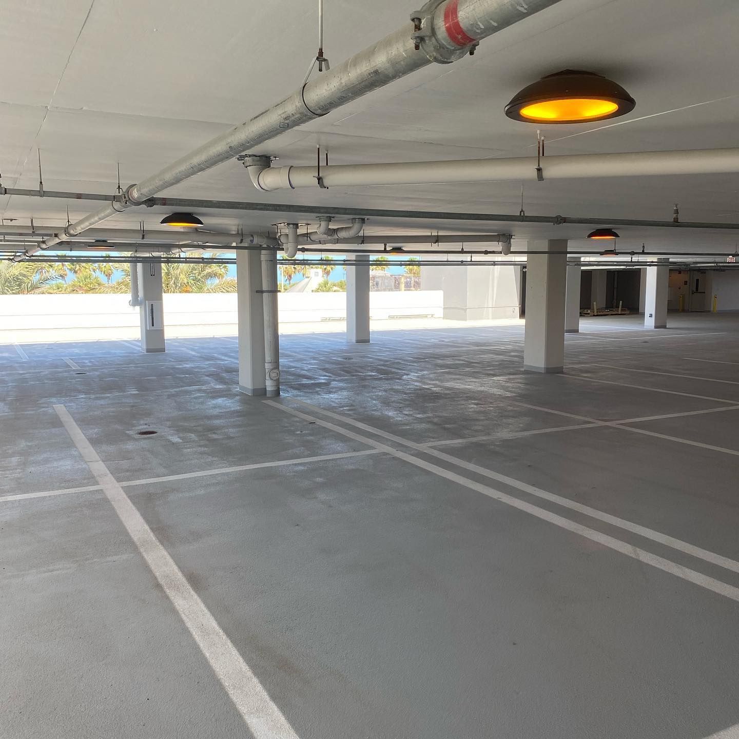 Empty, gray parking garage with white parking lines, concrete pillars, overhead pipes, and yellow lights.