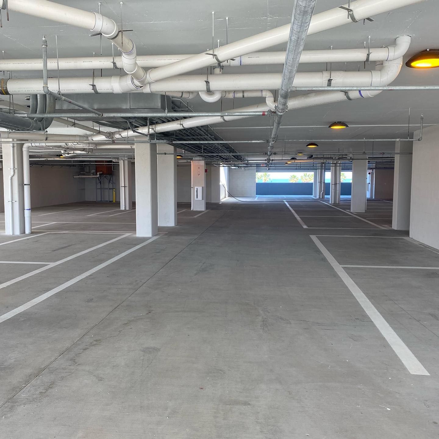 Empty parking garage with white pillars, lines, and overhead pipes; bright lighting.