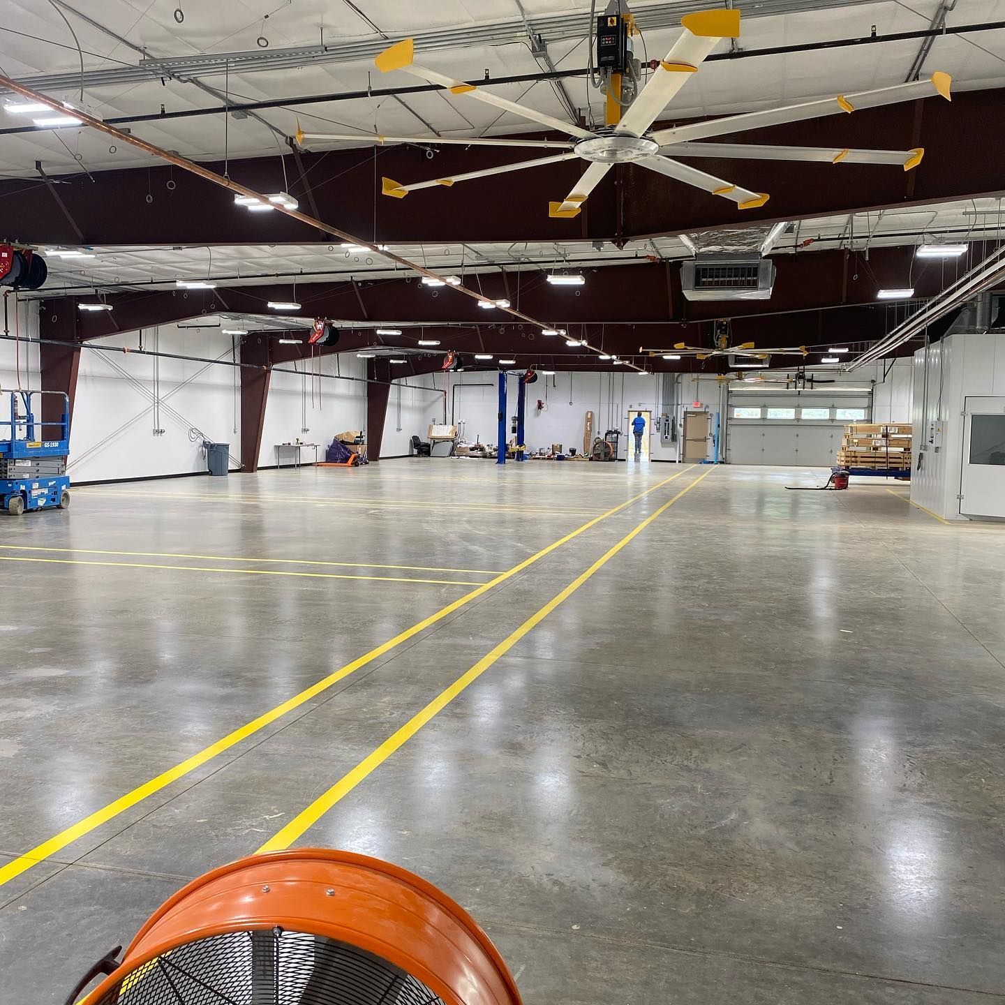 Large warehouse interior with high ceilings, concrete floor, and industrial fan in foreground. Yellow lines on floor.