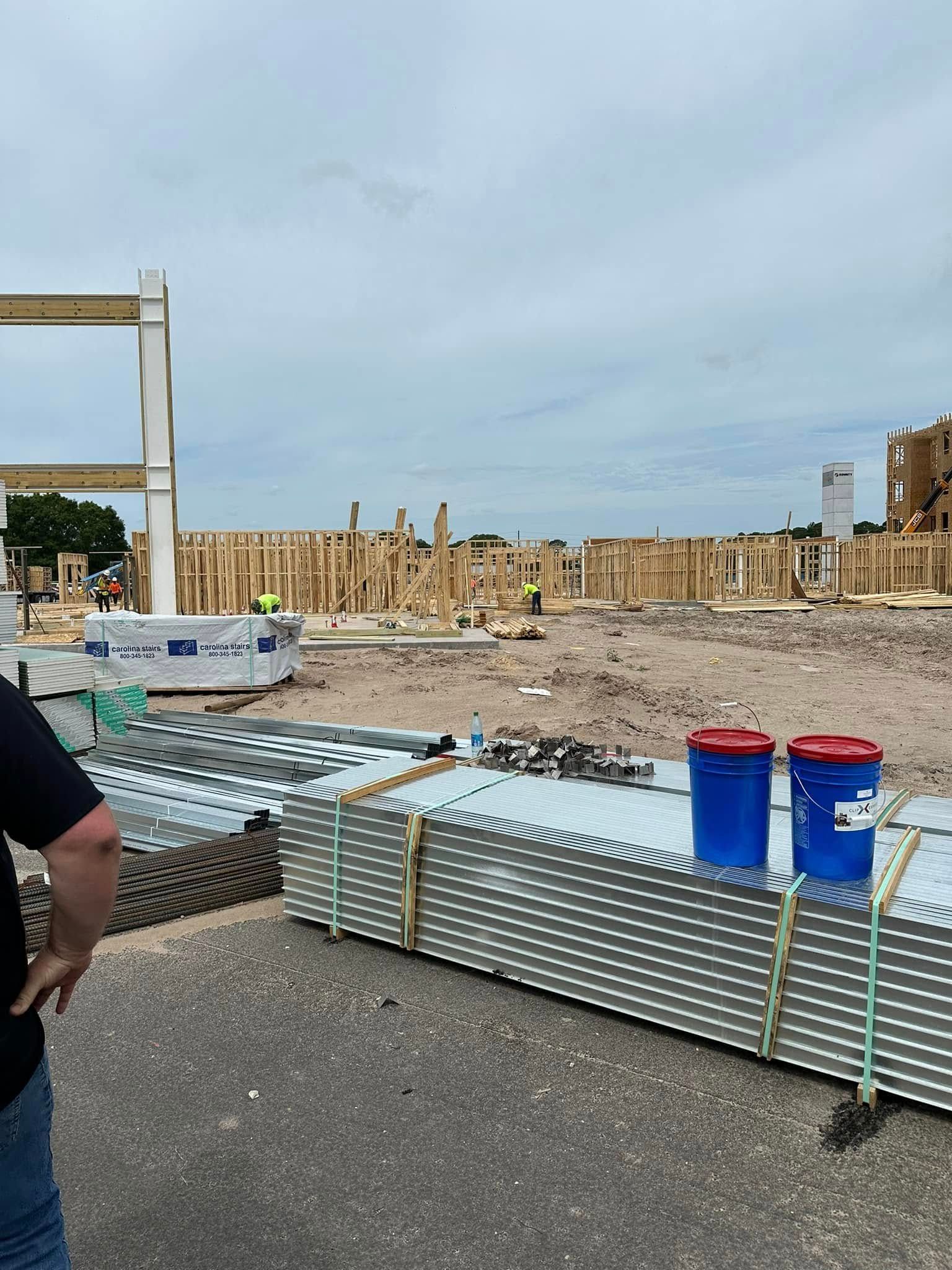 Construction site with exposed wooden framing and steel beams, overcast sky. Blue buckets sit on metal sheets.