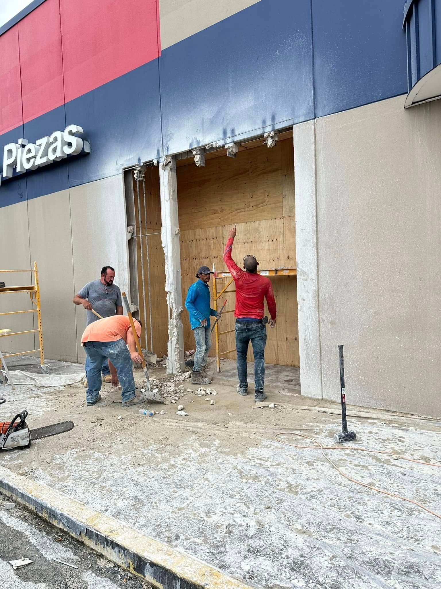 Workers at a store, ripping a section from the building. One points up; others work with tools on the ground.