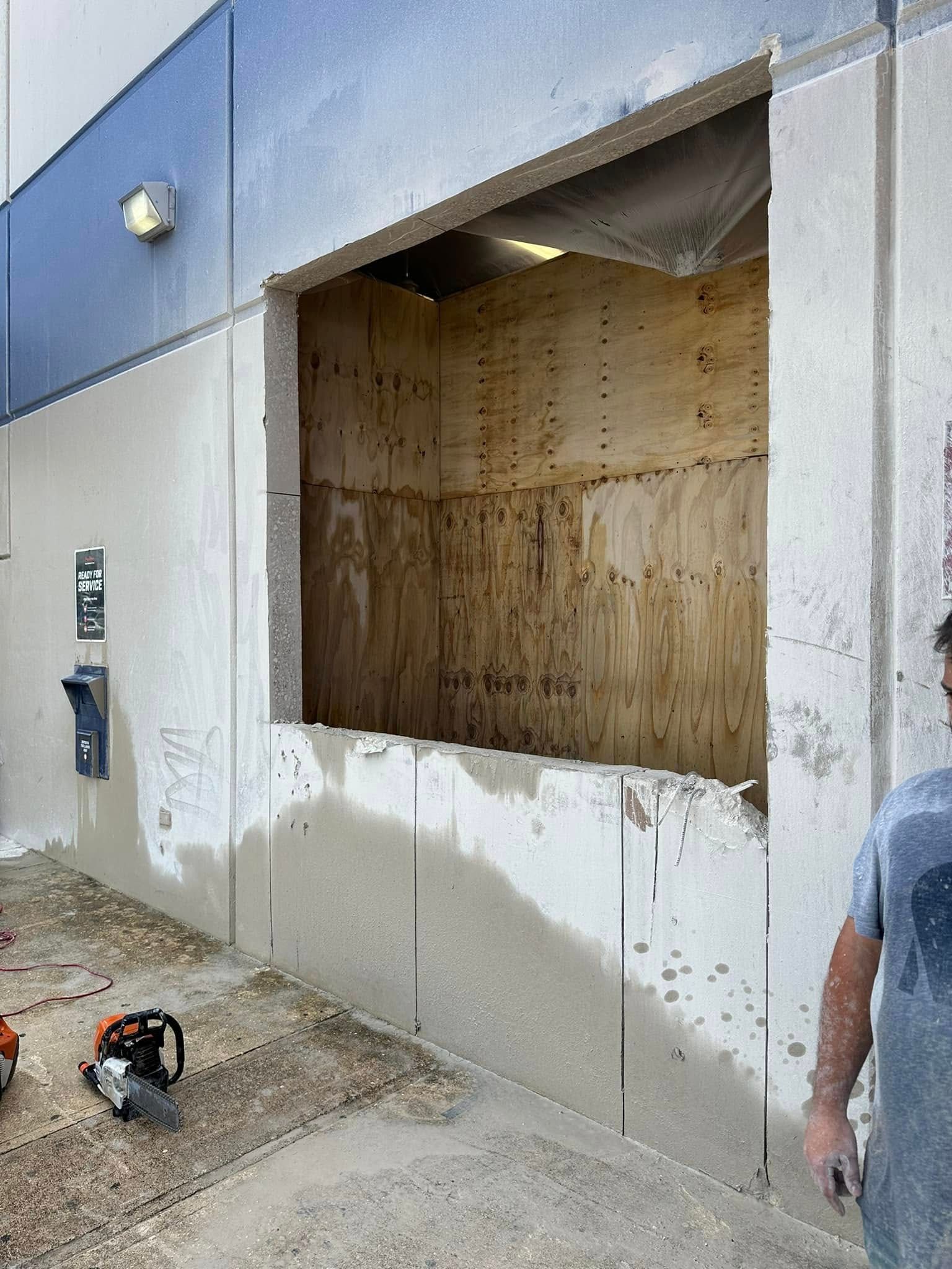 Man cutting a large rectangular opening in a concrete wall. Saw on ground. Building exterior.