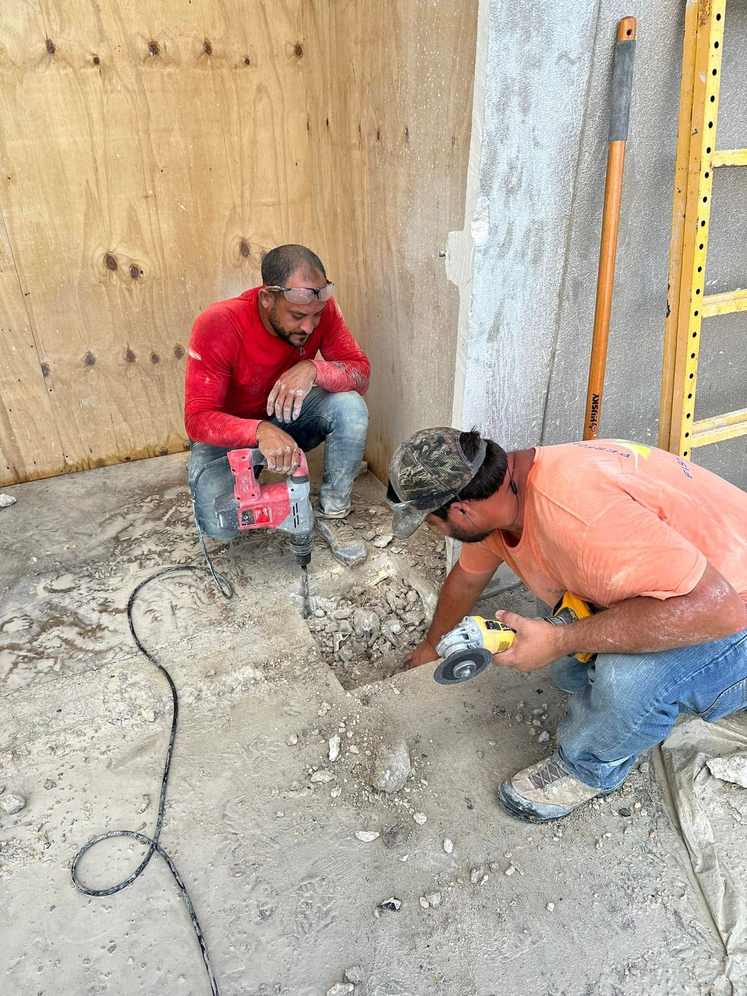 Two construction workers are breaking concrete. One kneels with a jackhammer, the other uses a grinder. Dusty environment.