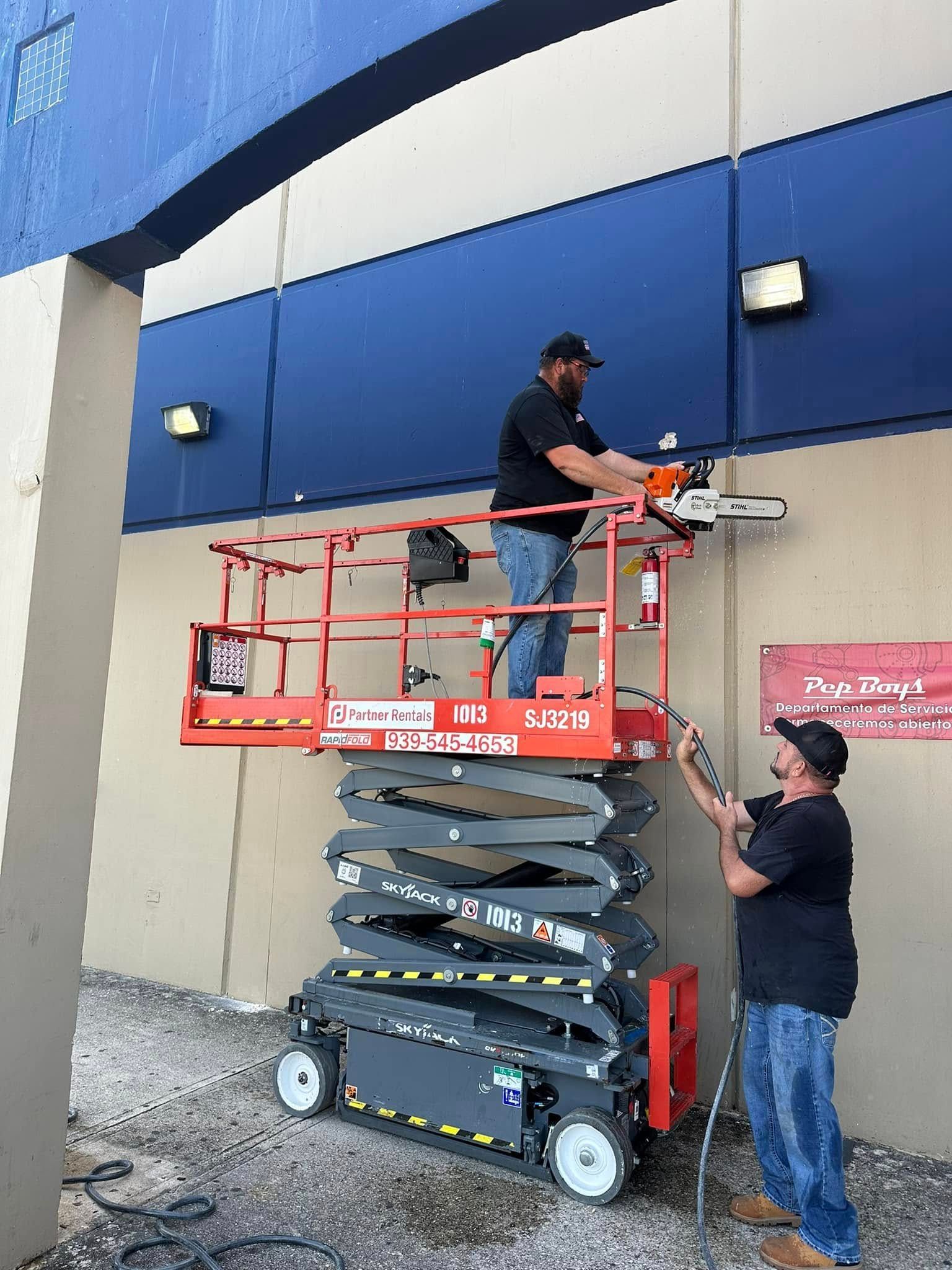 Two men using a lift with a chainsaw to work on a building exterior; one man operates the saw while the other works below.