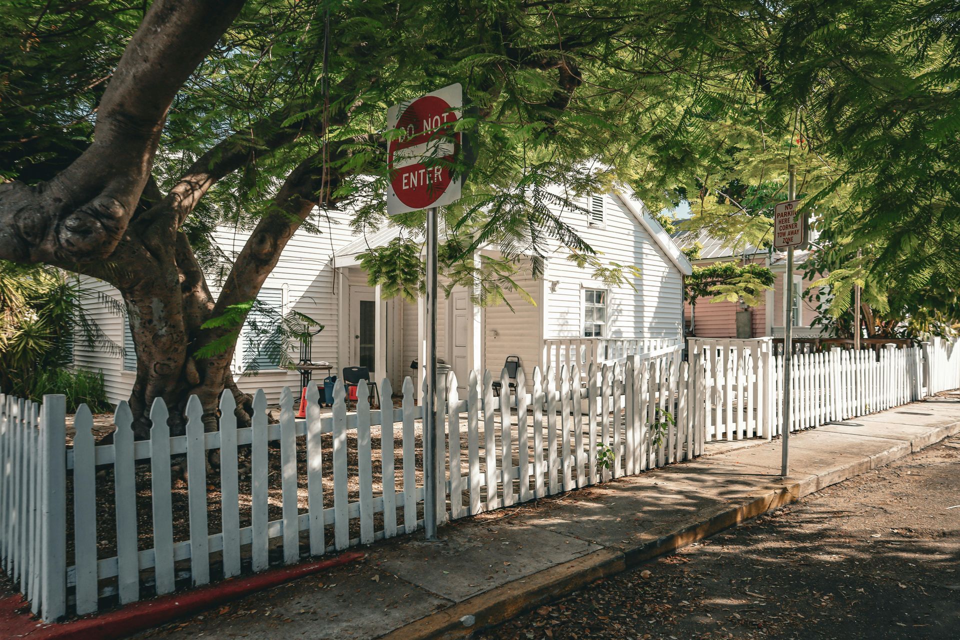 White picket fence around a house under leafy trees, with a stop sign beside the sidewalk.