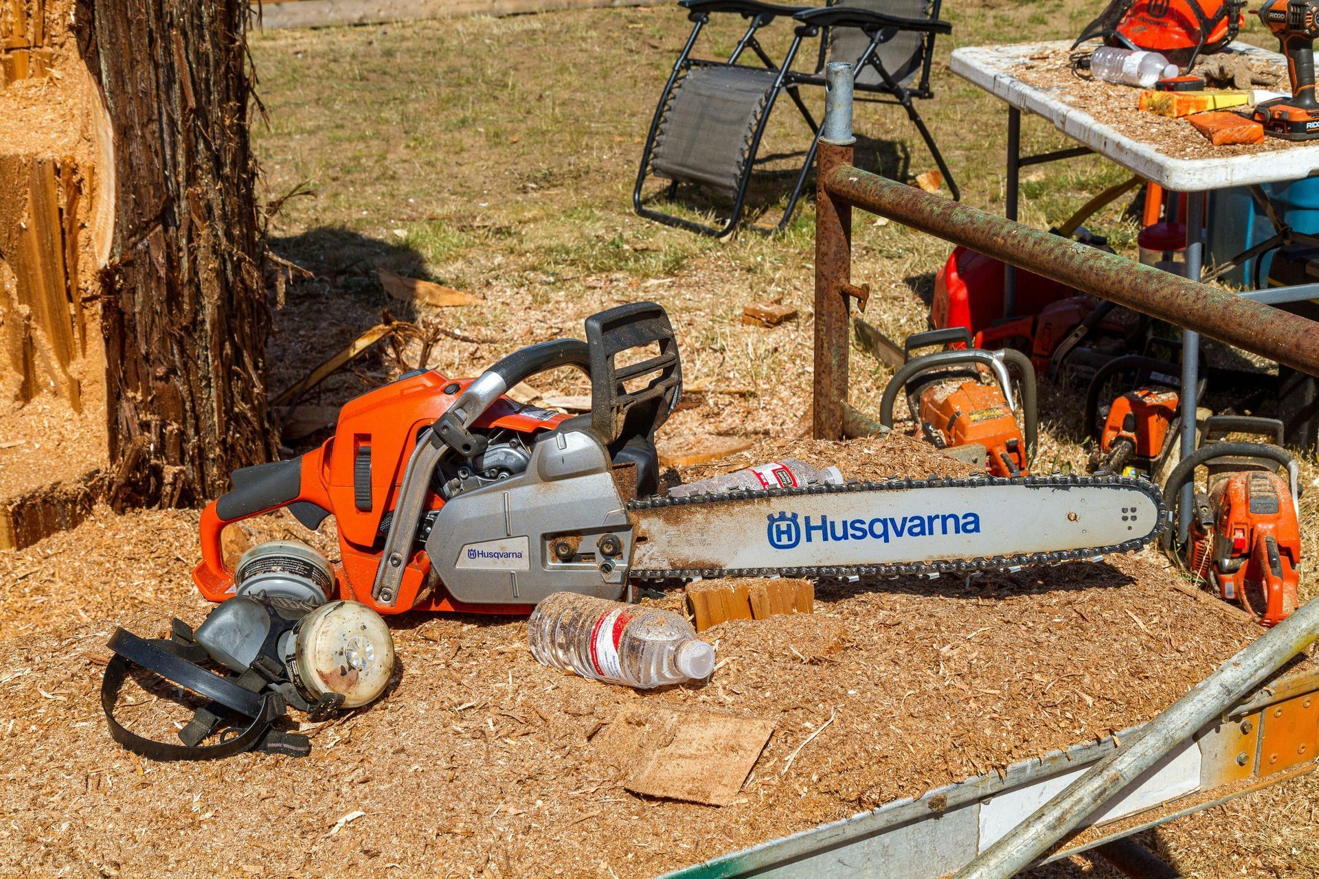 Chainsaw, helmet, and safety gear scattered on wood chips beside a tree and workbench