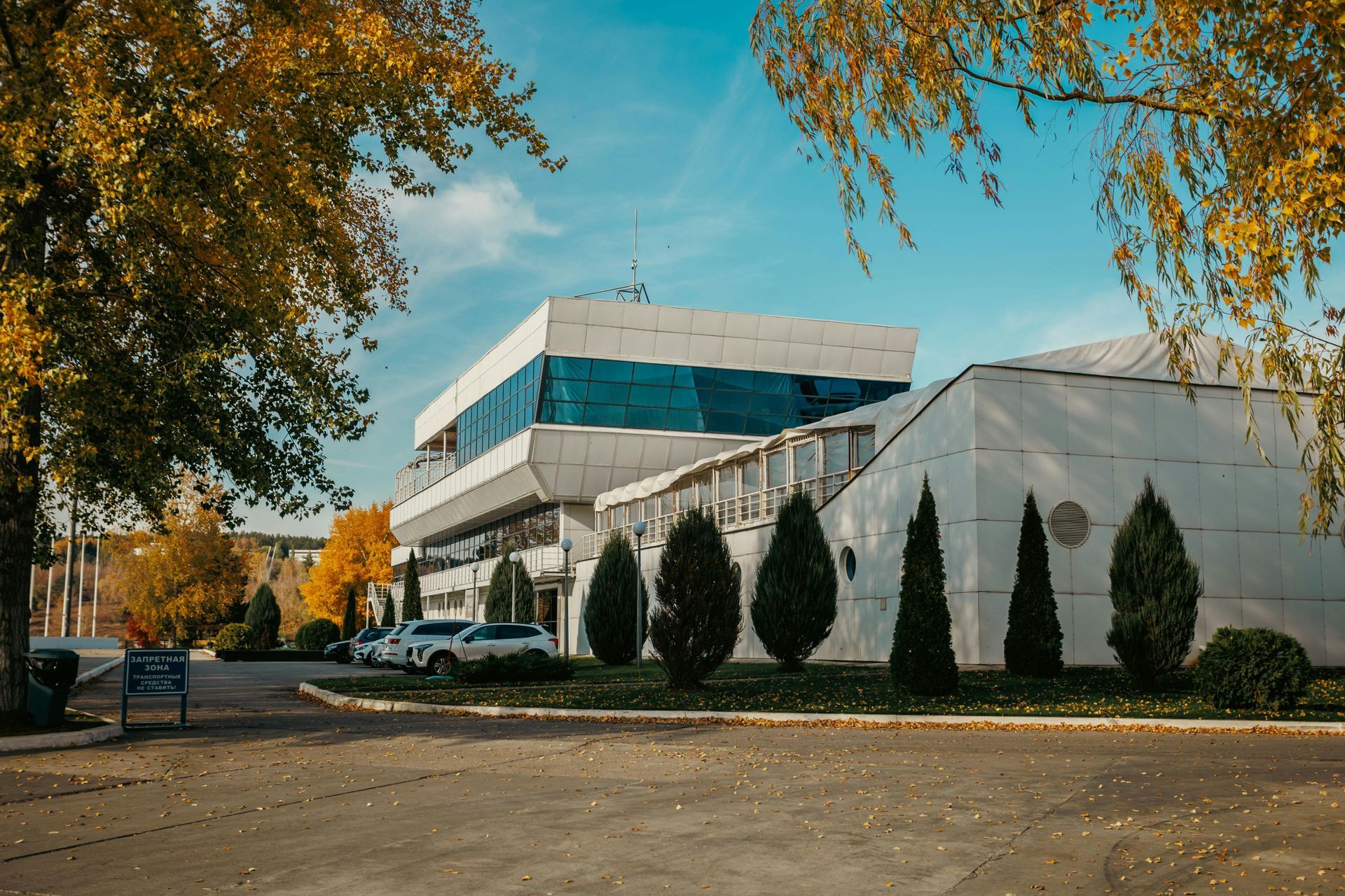 Low-rise office building with teal windows, evergreen shrubs, and autumn trees along a quiet street