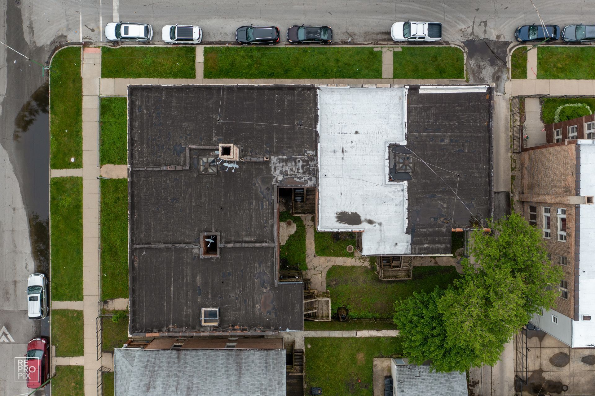 Overhead view of buildings with dark roofs, green lawns, and cars parked on street.