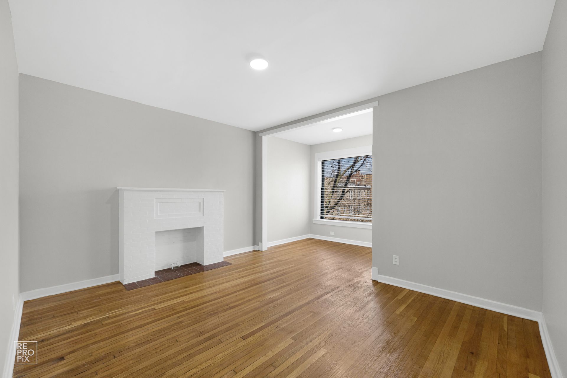 Empty room with hardwood floors, a white fireplace, and a window.