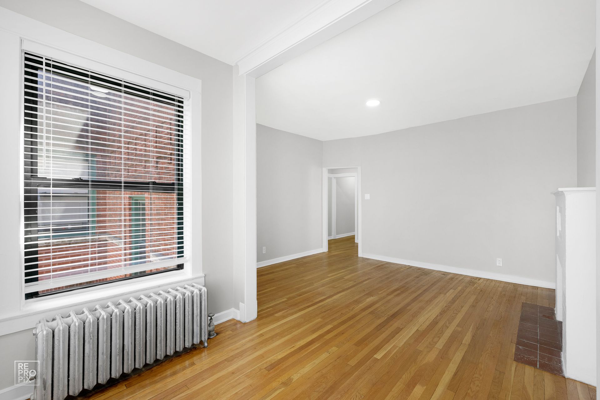 Empty living room with wood floors, large window with blinds, and radiator.