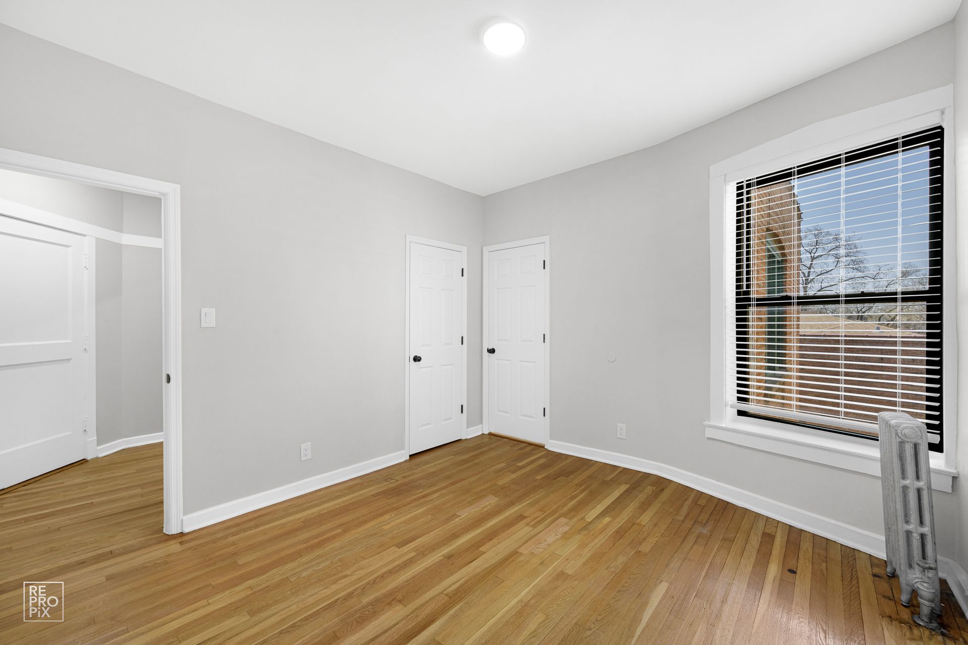 Empty bedroom with wood floors, two doors, and a window with blinds.