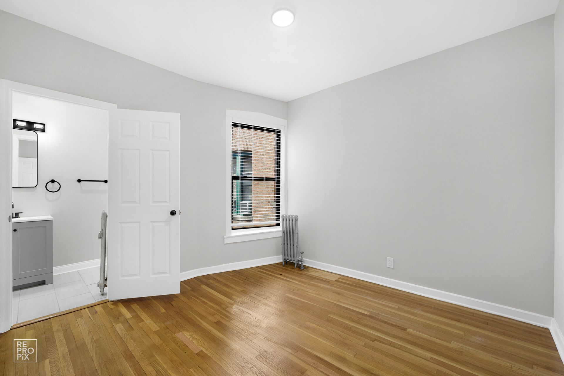 Empty room with hardwood floors, a bathroom, and a window with a radiator. Light gray walls.