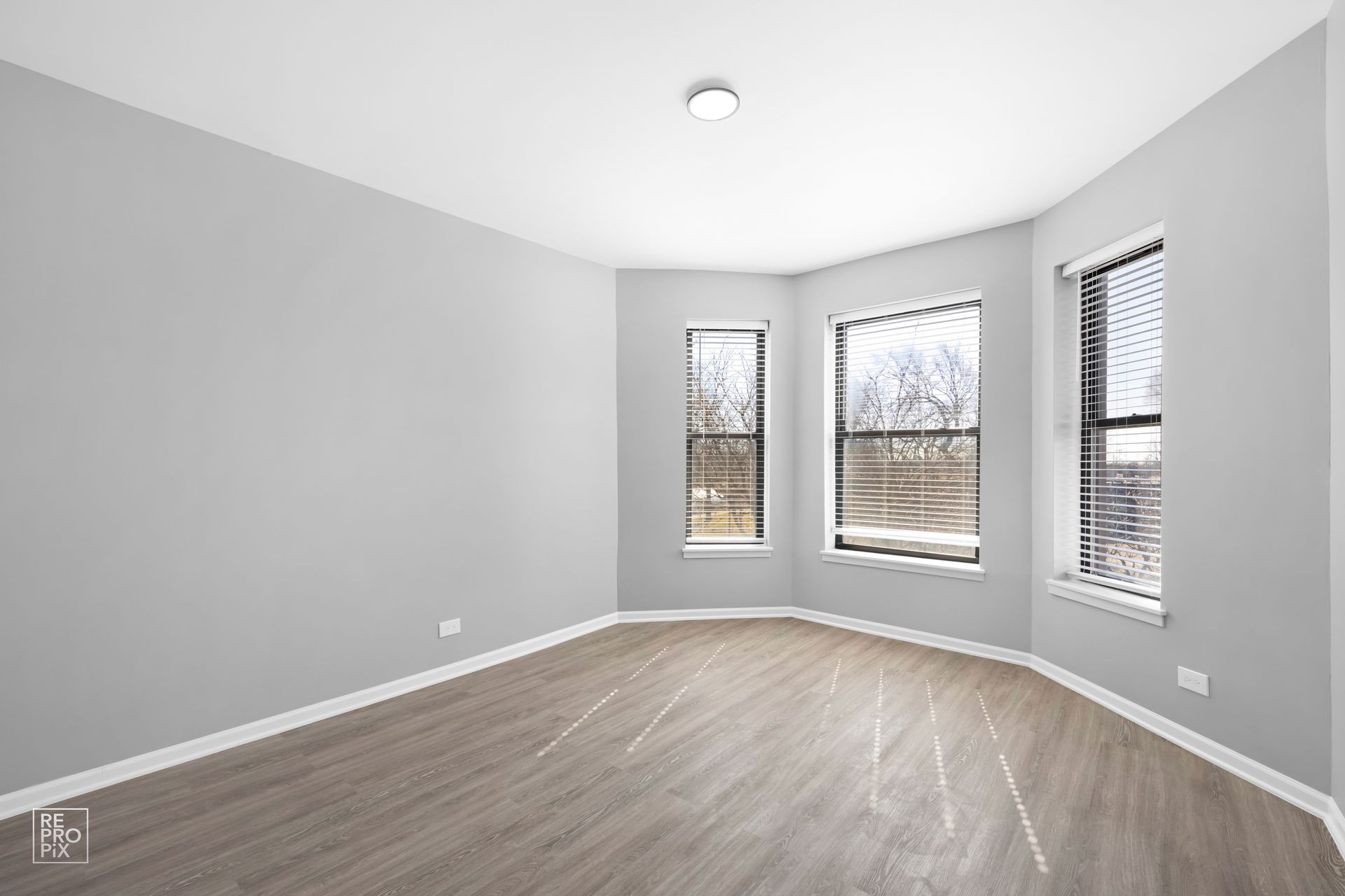 Empty room with grey walls, three windows with blinds, and wood-look flooring.