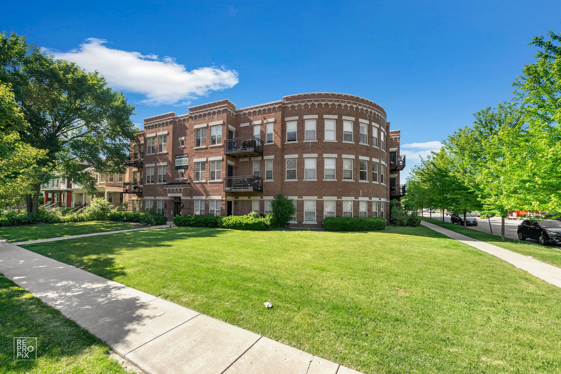 Three-story brick apartment building with a rounded facade, a grassy lawn, and sidewalk under a blue sky.