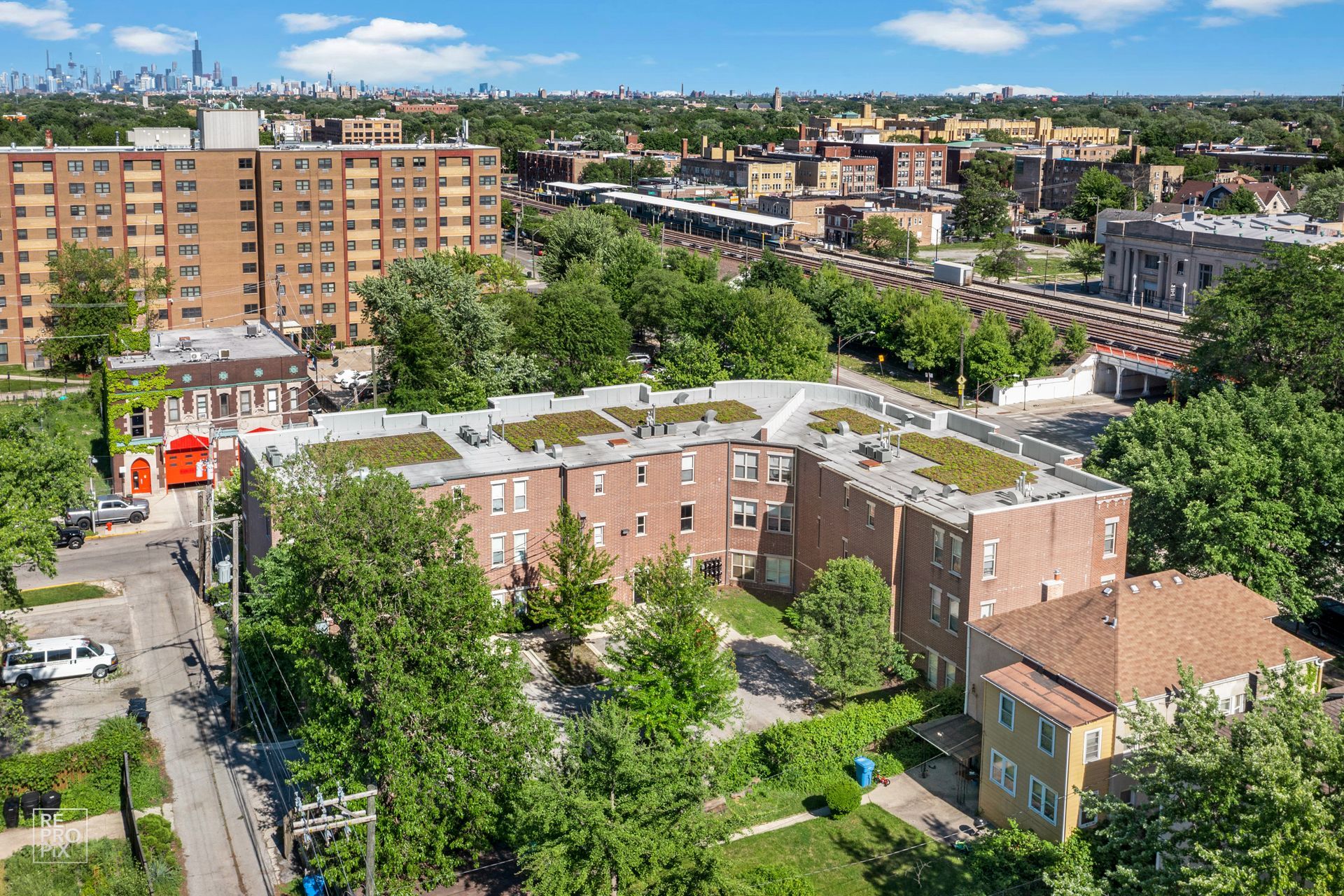 Aerial view of a multi-story brick apartment building with a green roof, surrounded by trees and other buildings.